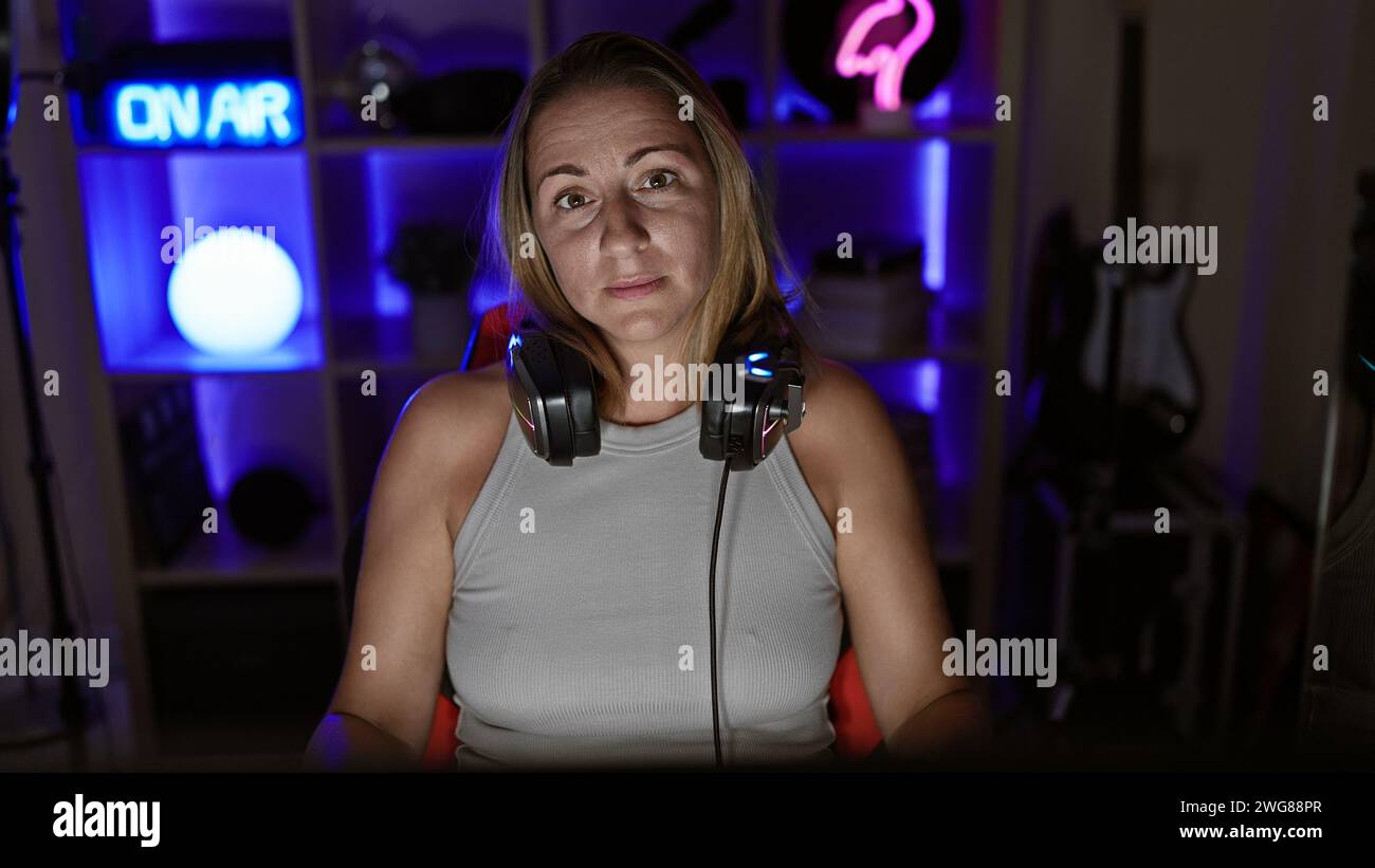 Beautiful, young, blonde female gamer and streamer, earnestly engaged in a nighttime gaming stream, seated in a tech-packed gaming room, geared with h Stock Photo