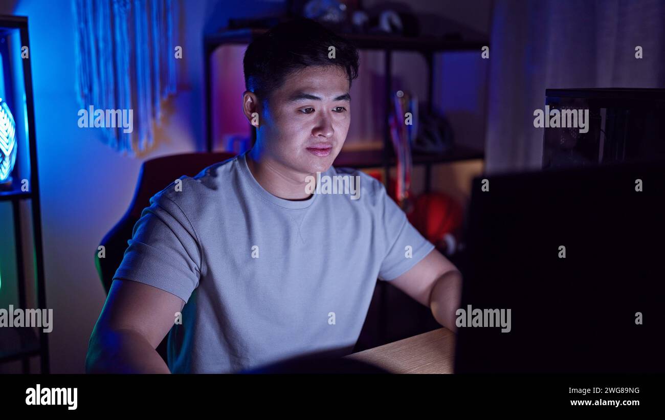 A young asian man focused while using a computer in a dark room with ambient lighting. Stock Photo