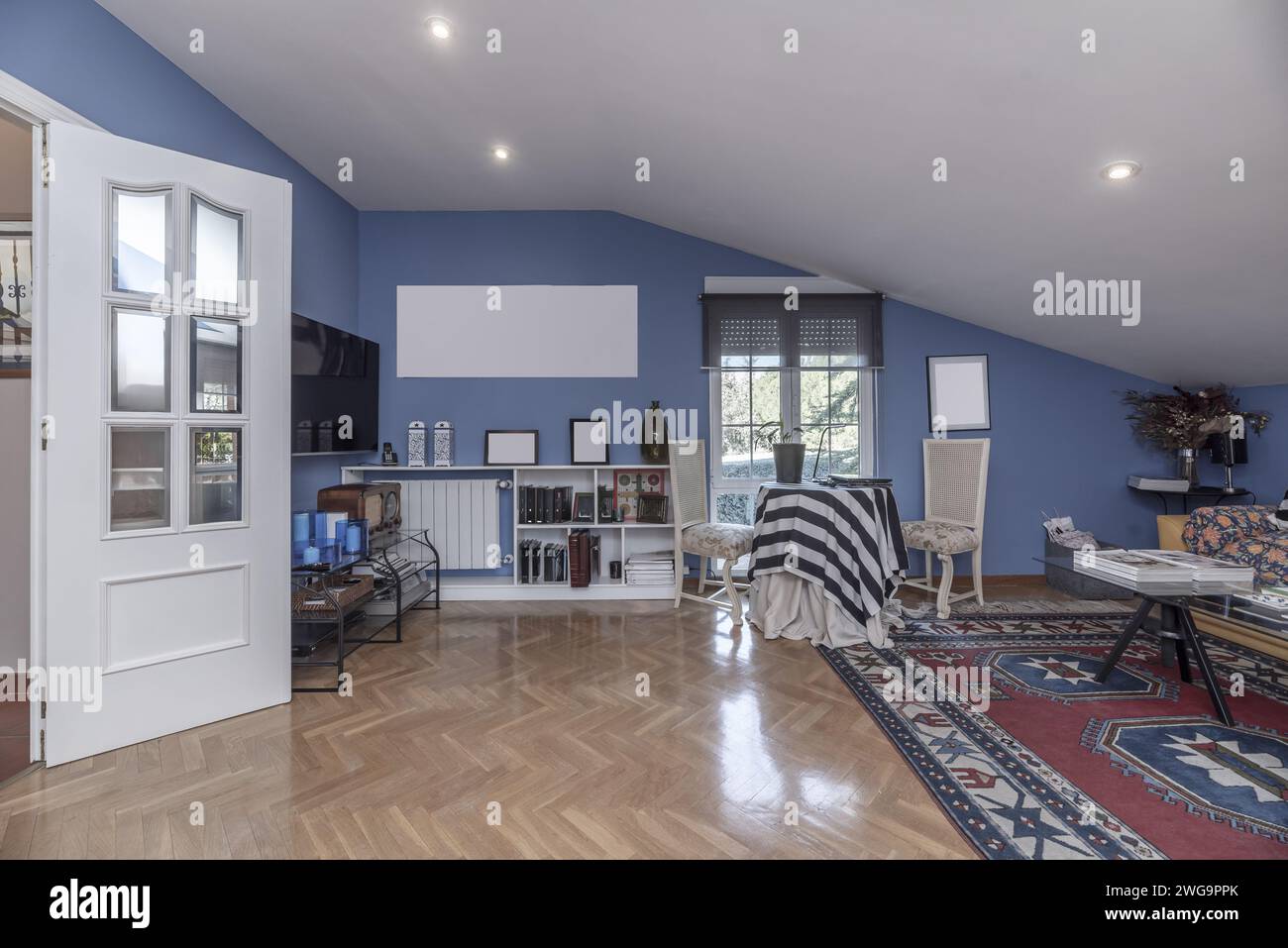 living room of a house with blue painted walls, white carpentry on the doors, sloping ceilings, oak parquet floors and carpets covering half Stock Photo