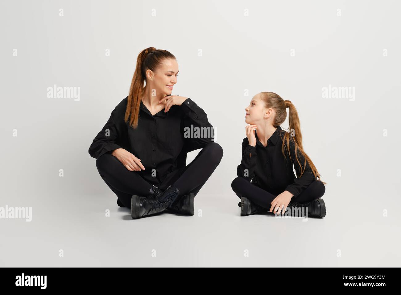 Mother and her daughter are sitting on a white background wearing similar stylish black shirt, tights and boots. They look at each other with smile Stock Photo