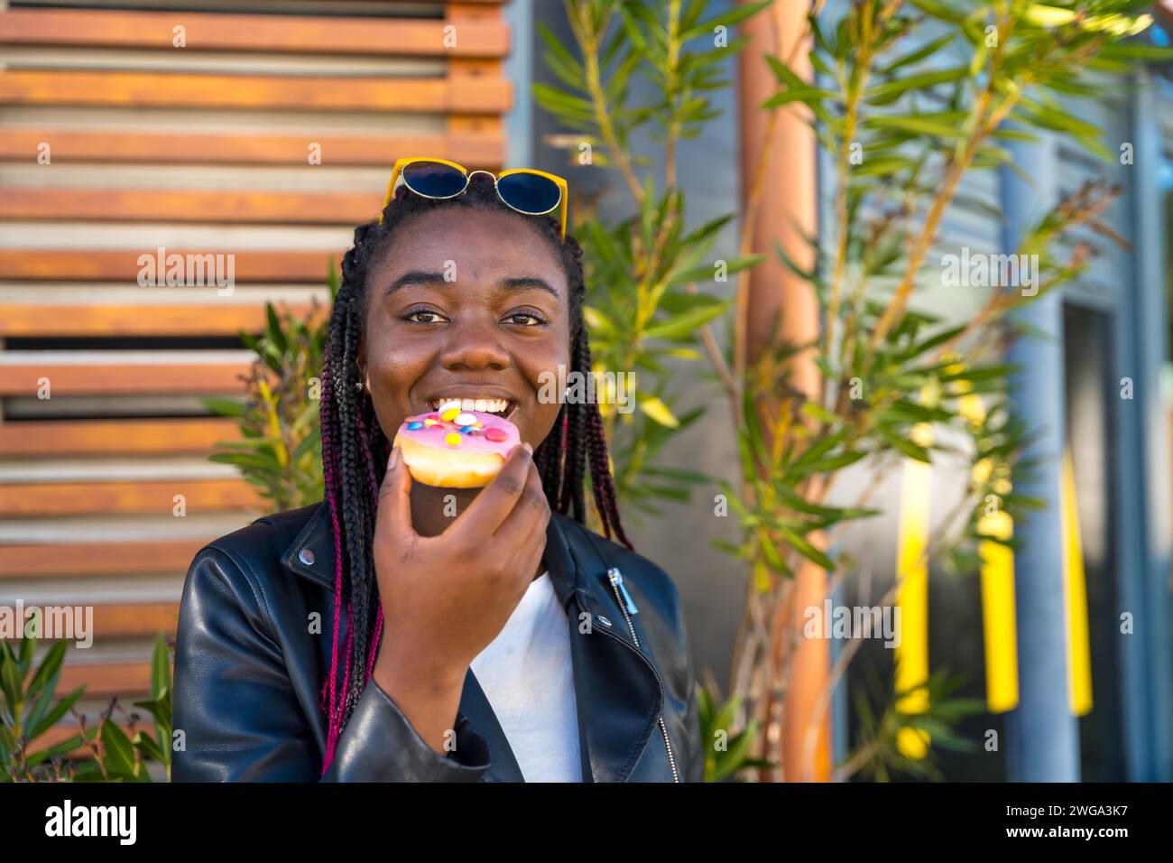 African cute woman eating a colorful doughnut sitting outdoors next to a shop Stock Photo