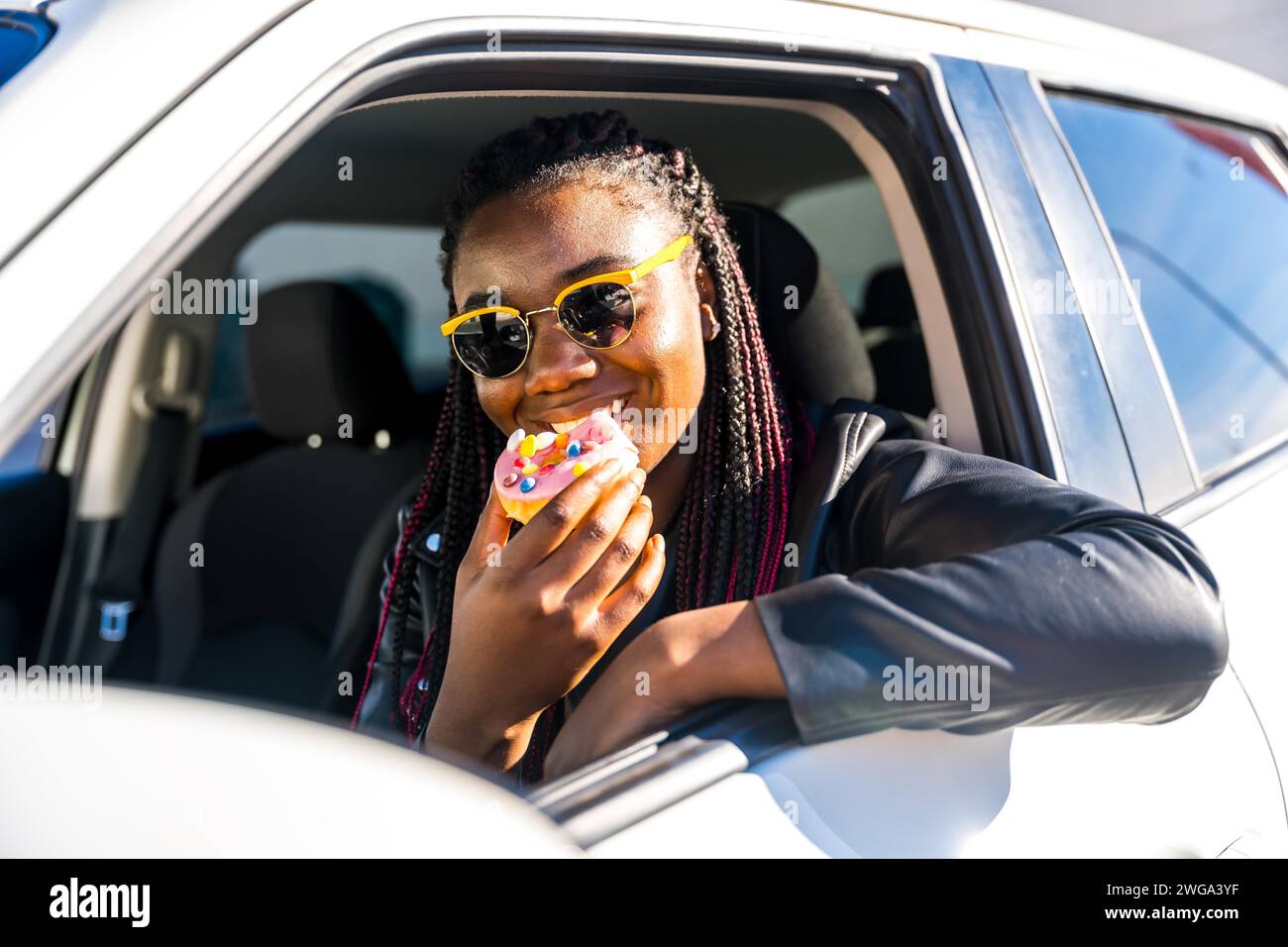 Cheerful african girl eating doughnut leaning in the car window sitting inside Stock Photo