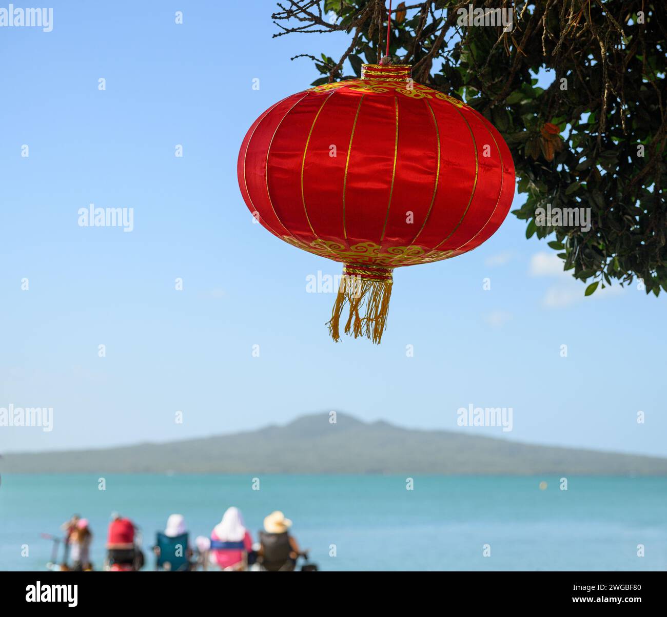 Chinese New Year lantern under the Pohutukawa tree. Unrecognizable people sunning and enjoying the view of Rangitoto Island. Mission Bay. Auckland. Stock Photo