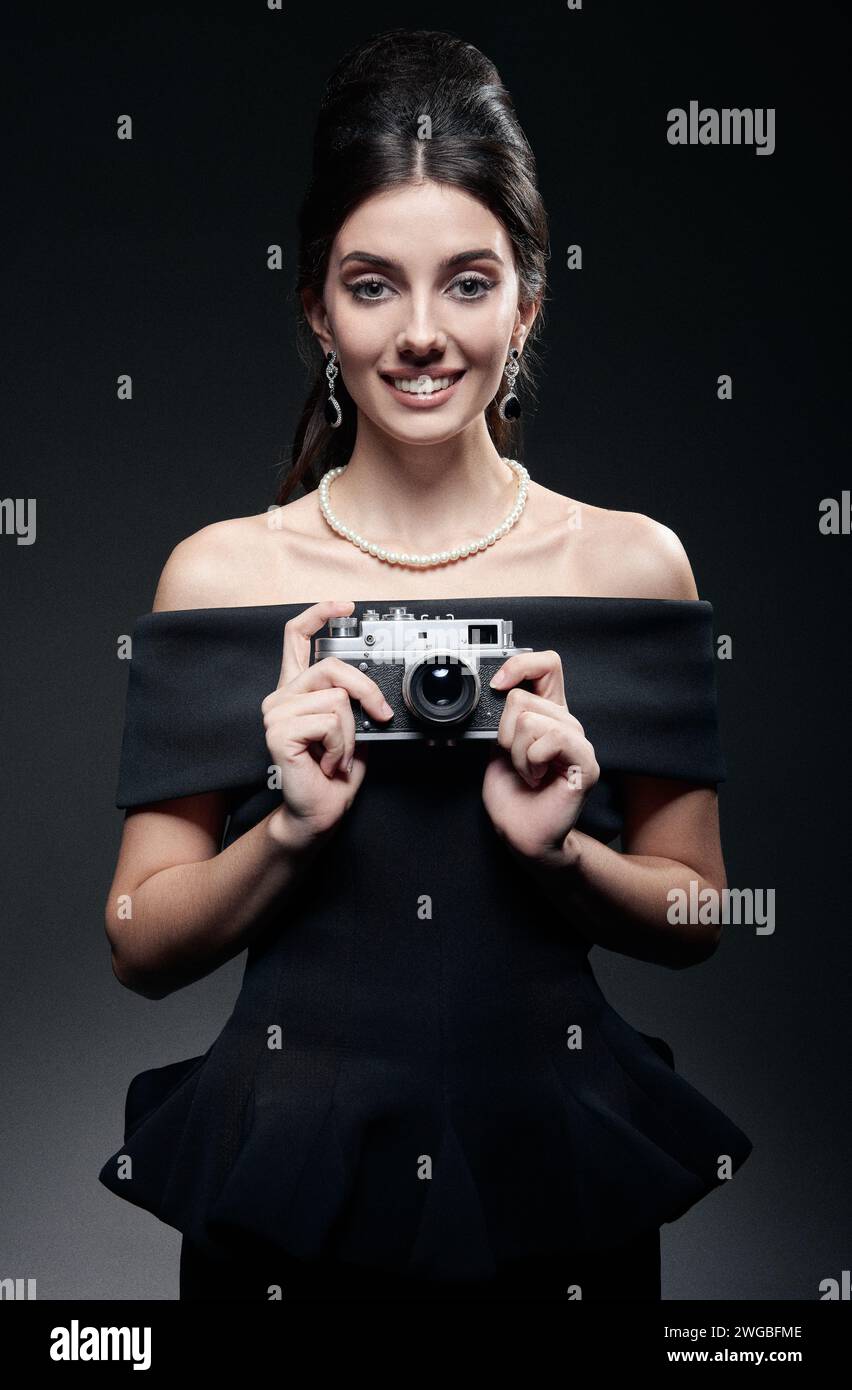 Retro shot: the beautiful young woman shooting by old film camera. Vintage portrait of smiling pretty girl in sixties style. Elegant lady in black dre Stock Photo