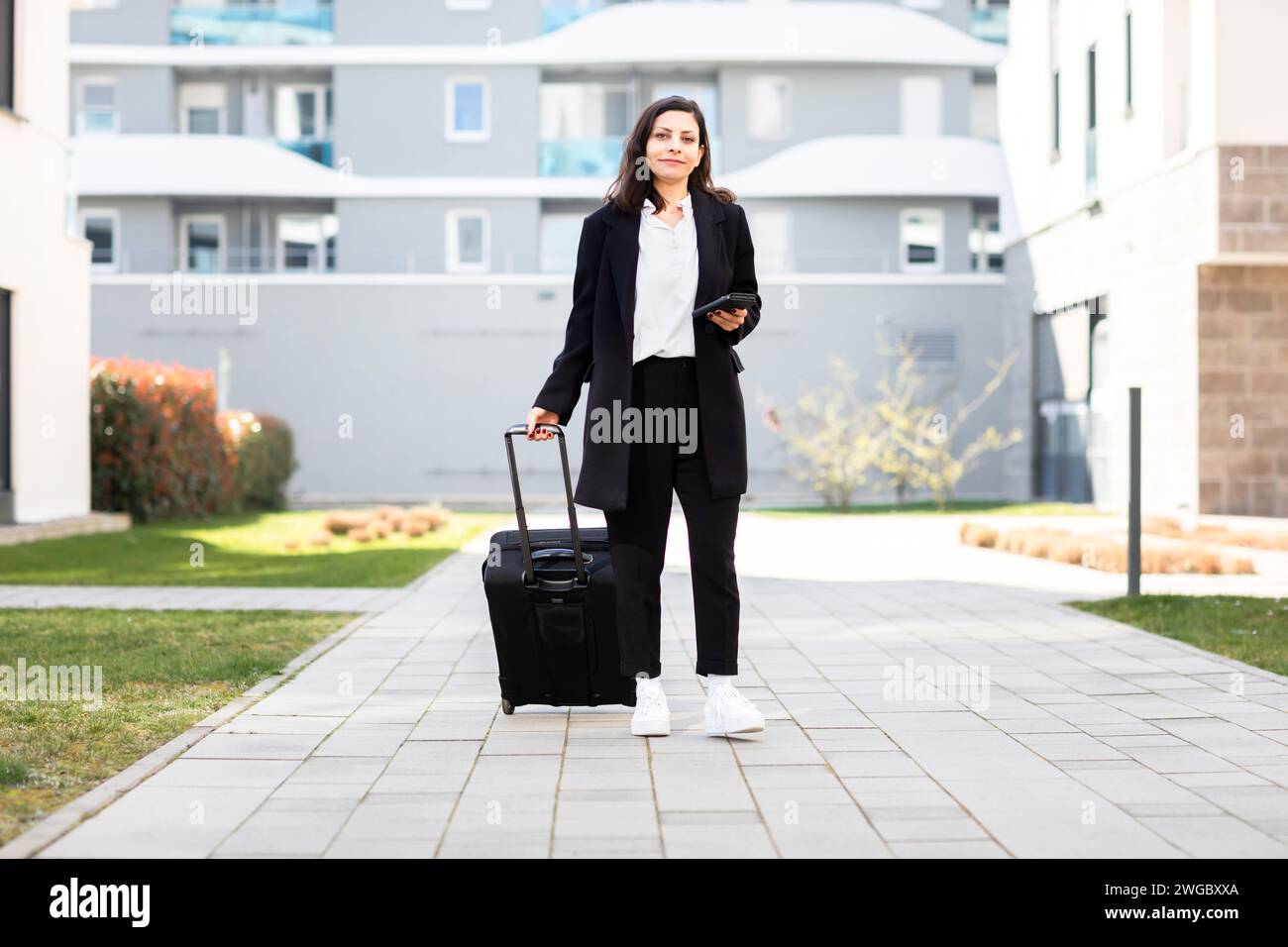 Woman standing in front of an apartment building with wheeled luggage, Germany Stock Photo