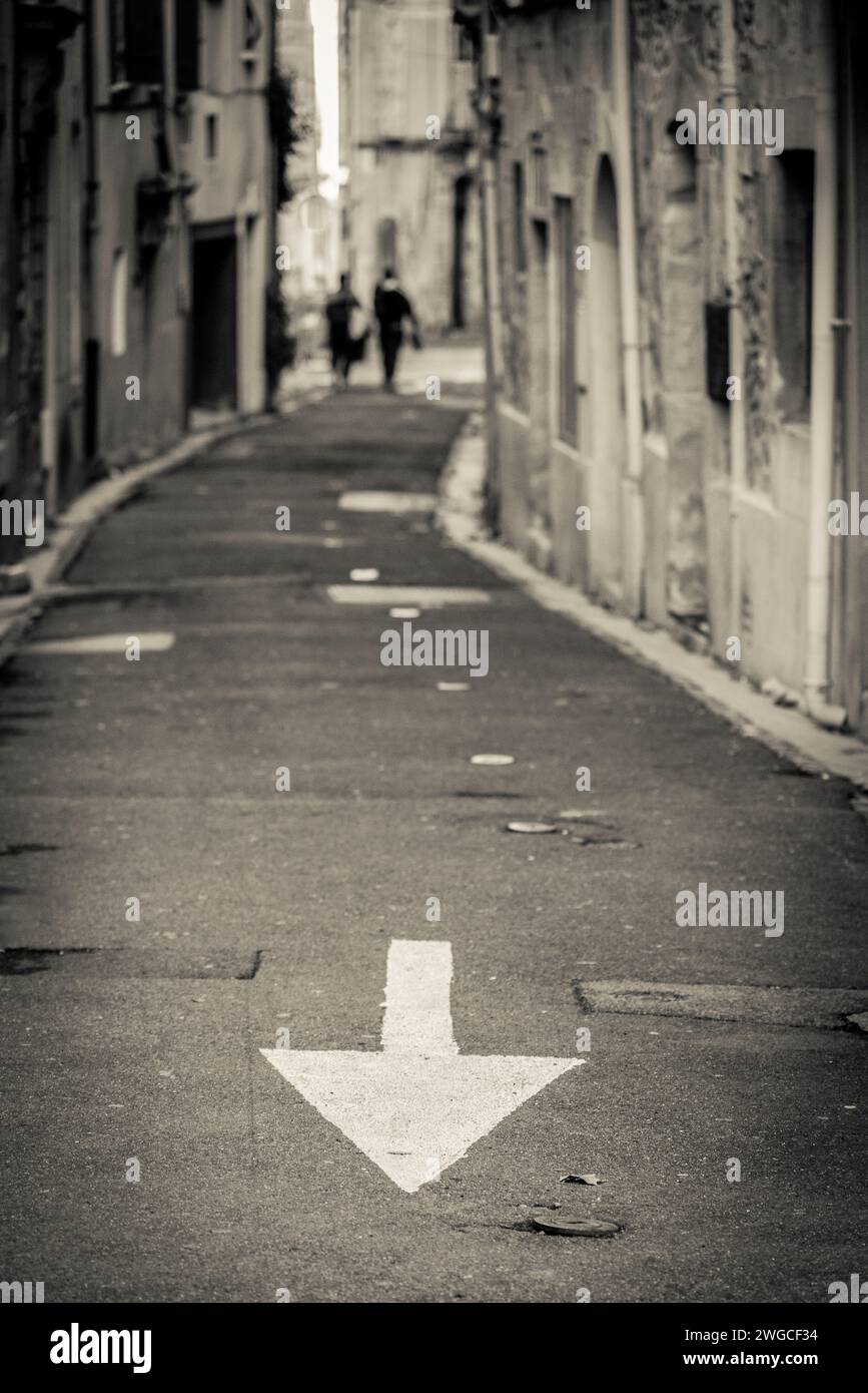 Two people walking away at the end of street and big traffic arrow pointing in opposite direction, Lodève, France Stock Photo
