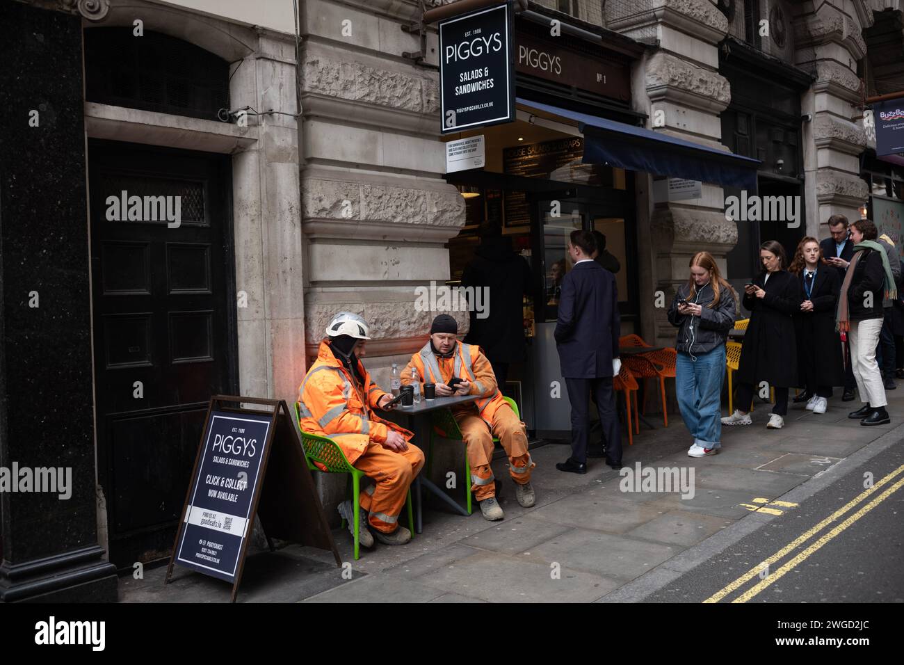 Manual workers sit outside at a sandwich bar table checking their smartphones whilst taking a coffee at lunchtime, London, England, UK Stock Photo