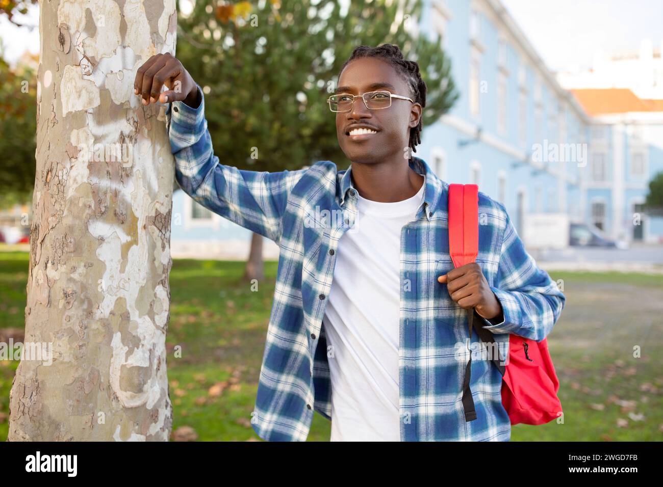 Cheerful black student guy with backpack leaning against tree outside Stock Photo