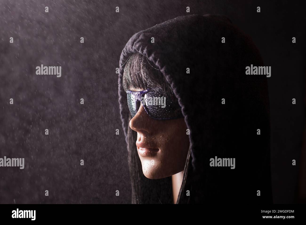 Plastic woman mannequin with black hair wearing a black hoodie and dark sunglasses posing on a black background in the mist Stock Photo