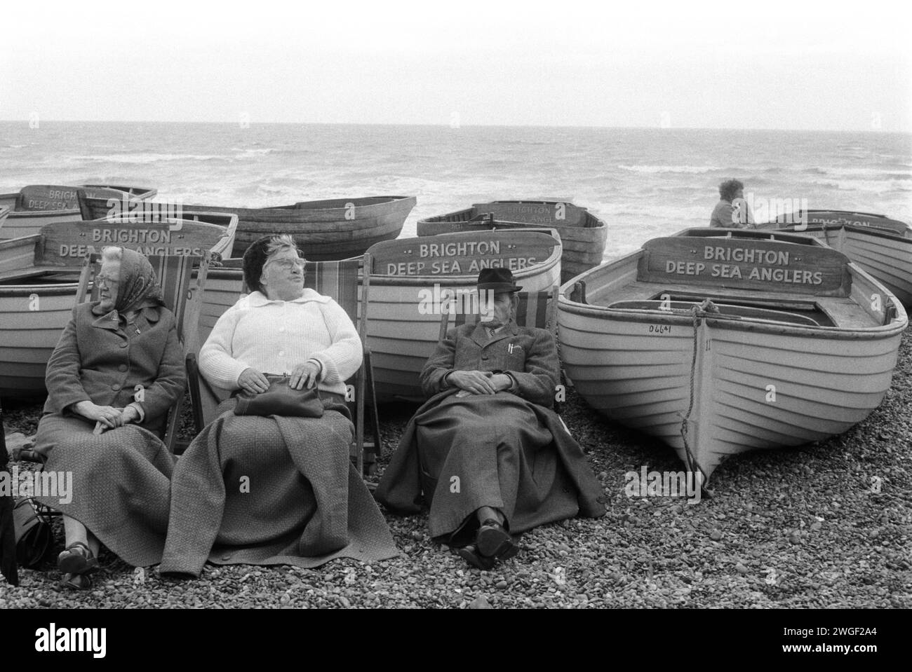 Brighton beach 1970s UK. Three older holidaymakers, wrapped in blankets sit on deckchairs keeping warm on Brighton beach, sheltering in part from the wind behind Deep Sea Anglers Club fishing boats. Brighton, East Sussex, England 1971. HOMER SYKES Stock Photo