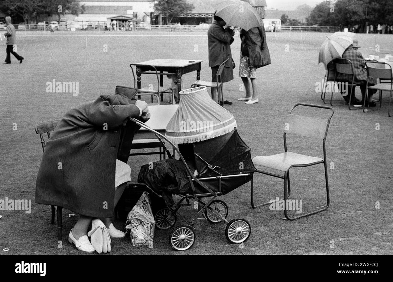 Mother with baby in pram sheltering from rain. 1970s UK. Church of St. John the Baptist annual summer church fete, women shelter from the rain. Cirencester, Gloucestershire. 1974 1970s UK HOMER SYKES Stock Photo