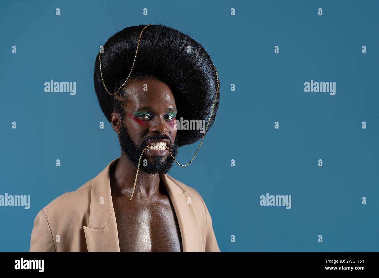 Portrait of African American gay man with makeup and black vintage wig. LGBT person holding gold accessory with teeth and wearing jacket on blue background. Stock Photo