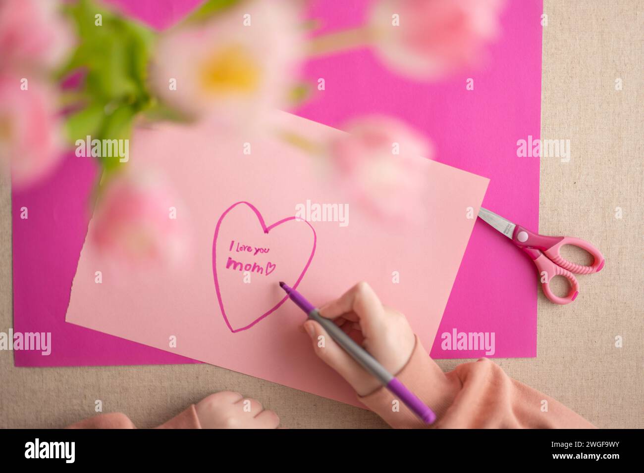 DIY mom card. Mothers Day.Child draws a heart on a pink piece of paper and writes mom I love you. daughter makes a card for his mother.Flowers and Stock Photo
