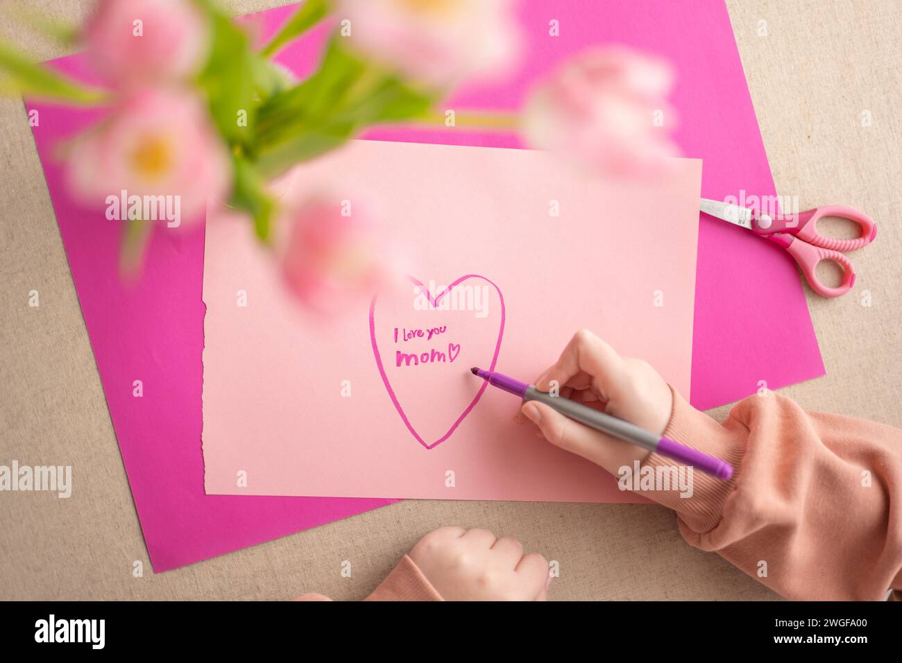 moms day. DIY mom card. Mothers Day.Child draws a heart on a pink piece of paper and writes mom I love you. daughter makes a card for his mother Stock Photo