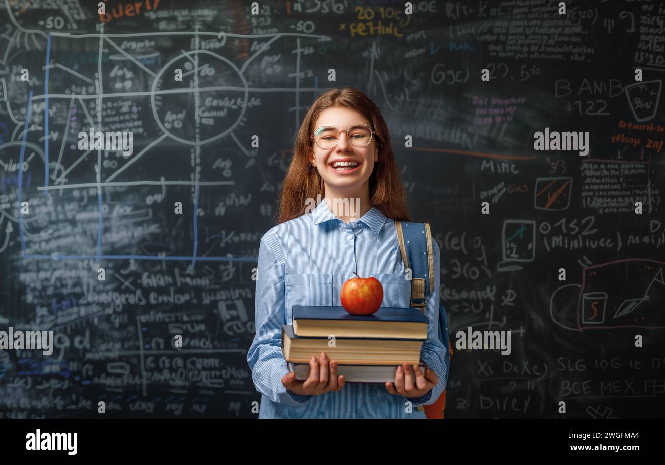 Back to school. Happy cute industrious teenager indoors. Girl is learning in class on background of blackboard. Stock Photo