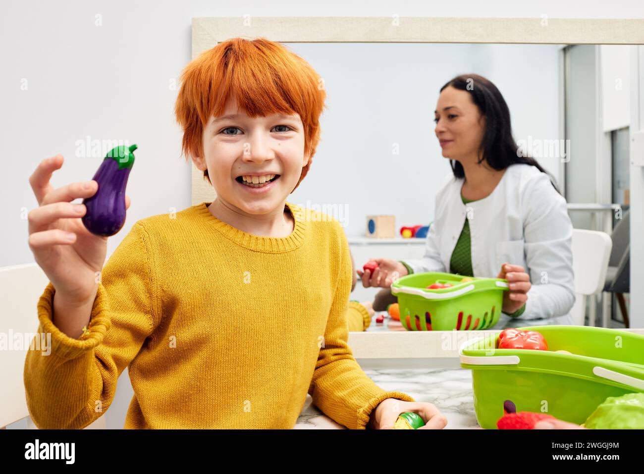 Red-haired boy holding toy eggplant during speech training session with his speech therapist. Children's speech and language therapy Stock Photo