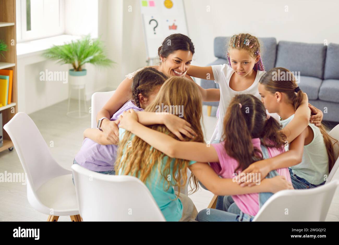 School girls sitting in a circle and hugging with psychologist woman during therapy session. Stock Photo