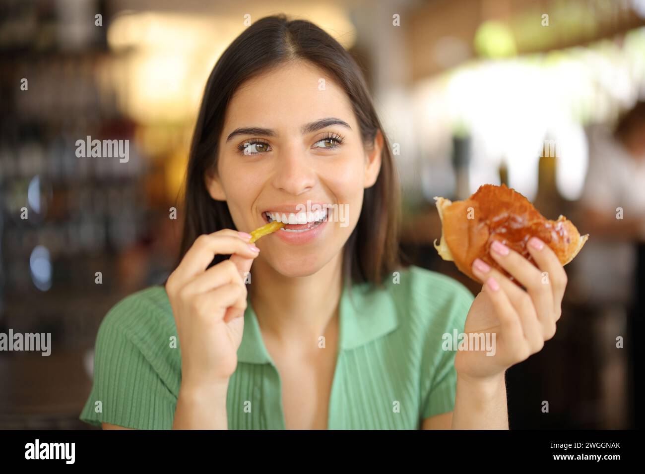 Happy woman eating burger and fries in a coffee shop looking at side Stock Photo