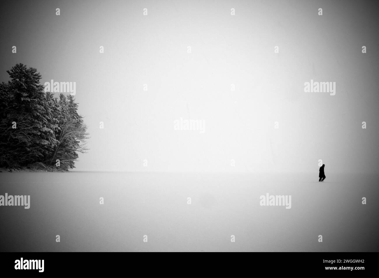 A man in a long black coat walks across a frozen and barren lake in a snowstorm Stock Photo