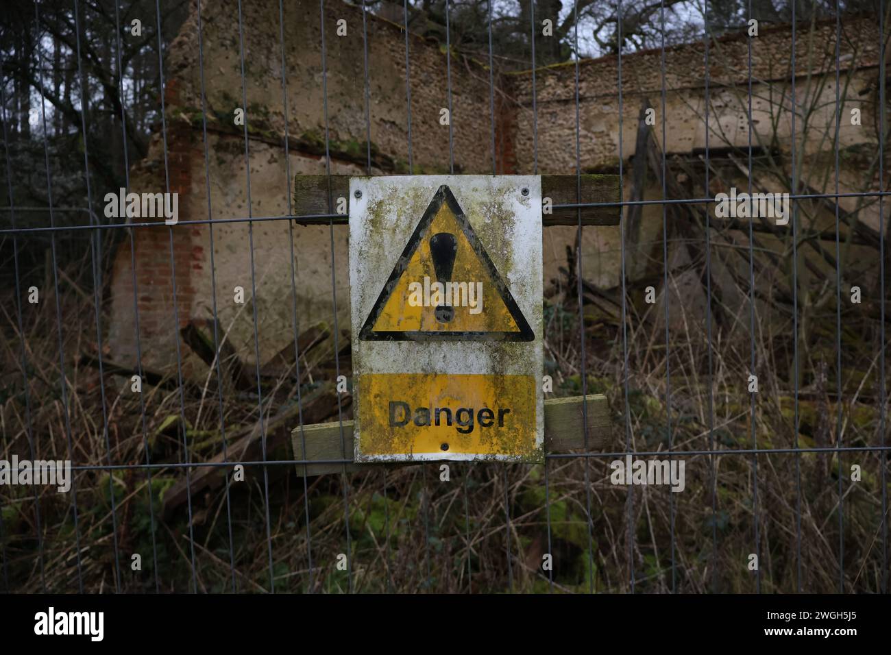 A dirty 'Danger' warning sign placed on a wire fence in front of a destroyed, abandoned house. There is debris and collapsed walls behind it. Stock Photo