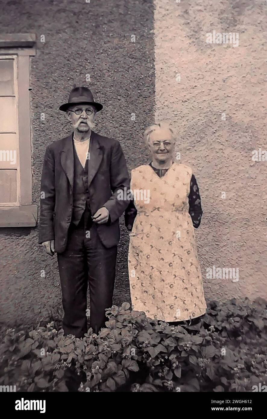 Taken from the original negative (so blemishes maybe present), a photo of a couple standing in their garden in the early 1940's. The man has a handlebar moustache and is wearing a trilby hat. The lady is wearing a long housecoat (pinafore). Stock Photo