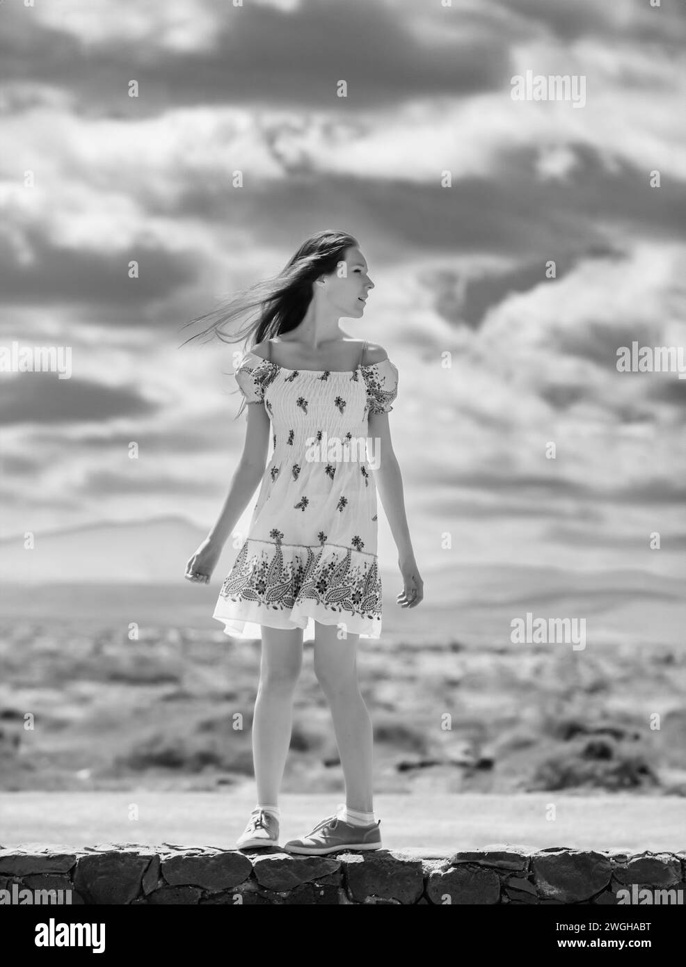 black and white photograph of a teenage girl with a white summer dress against a background of clouds Stock Photo