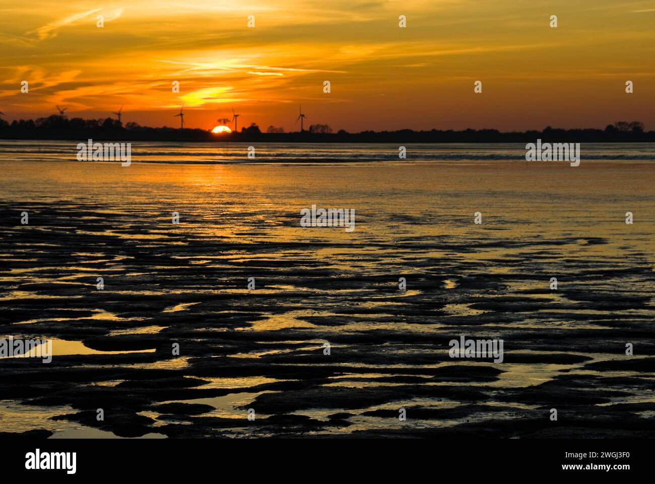 Vibrant sunrise over calm water and rocky shore, showcasing a distant sailboat Stock Photo