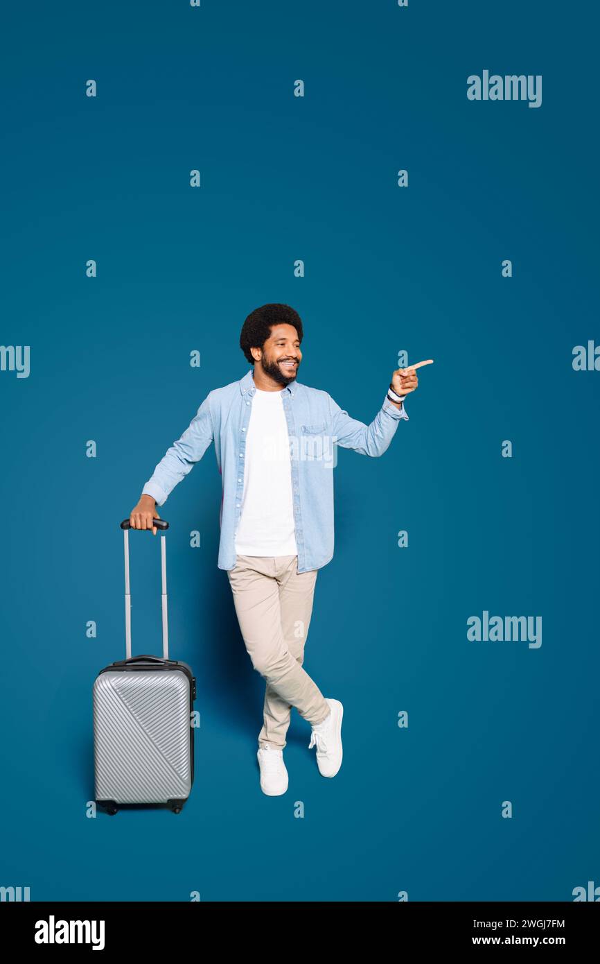 The jovial man standing with a suitcase, pointing aside, his face alight with enthusiasm, suggesting an inviting destination or a spontaneous travel opportunity, travel and holiday concept, copy space Stock Photo