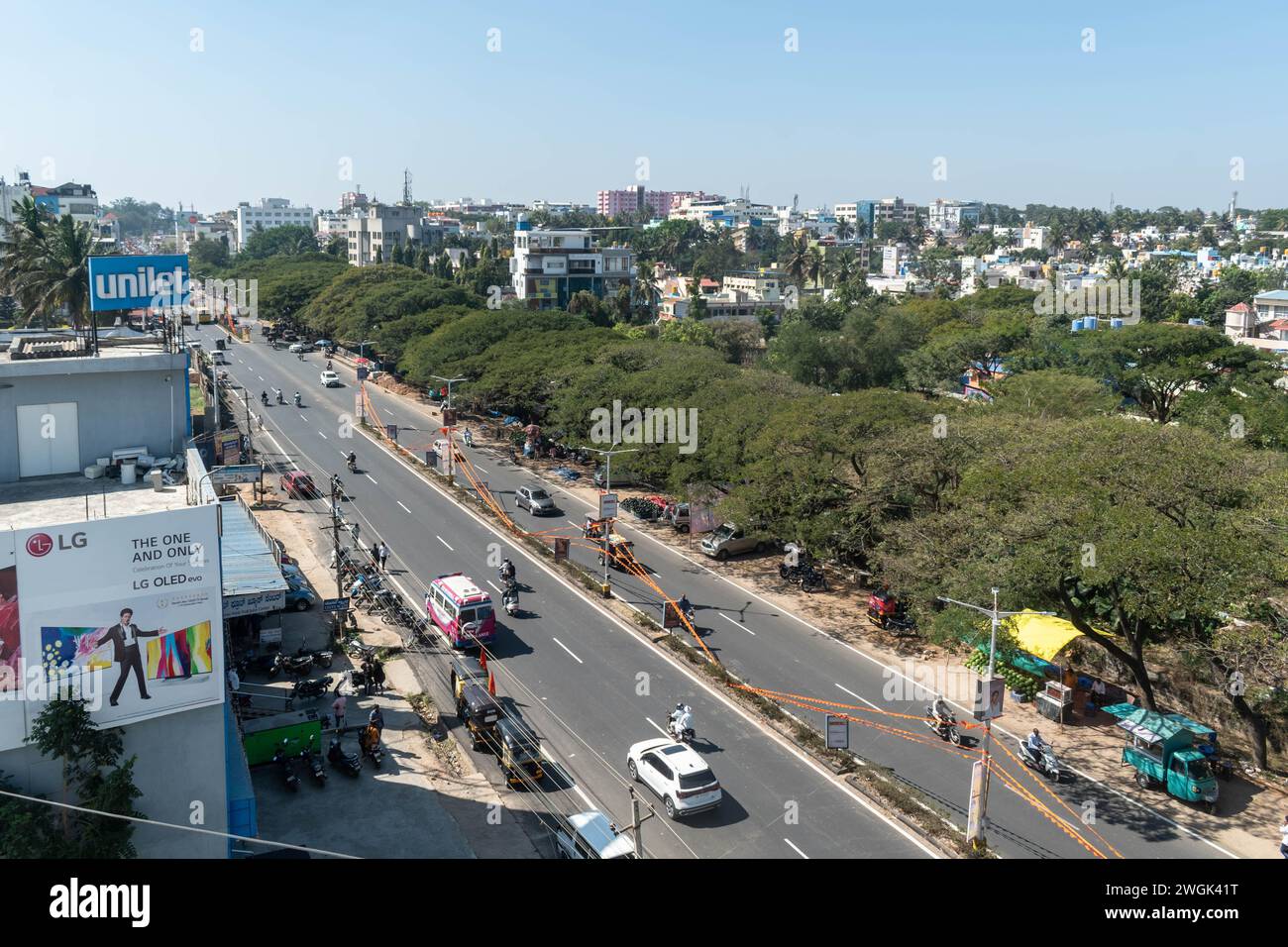 Hassan, Karnataka, India - January 10 2023: Clear sky over a bustling street, lined with shops and vehicles, showcasing urban life in a growing city d Stock Photo