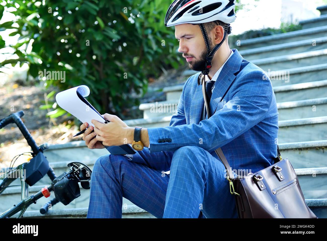 Young businessman in helmet sitting on stairs and analyzes business report in clipboard. Business and working concept. Stock Photo