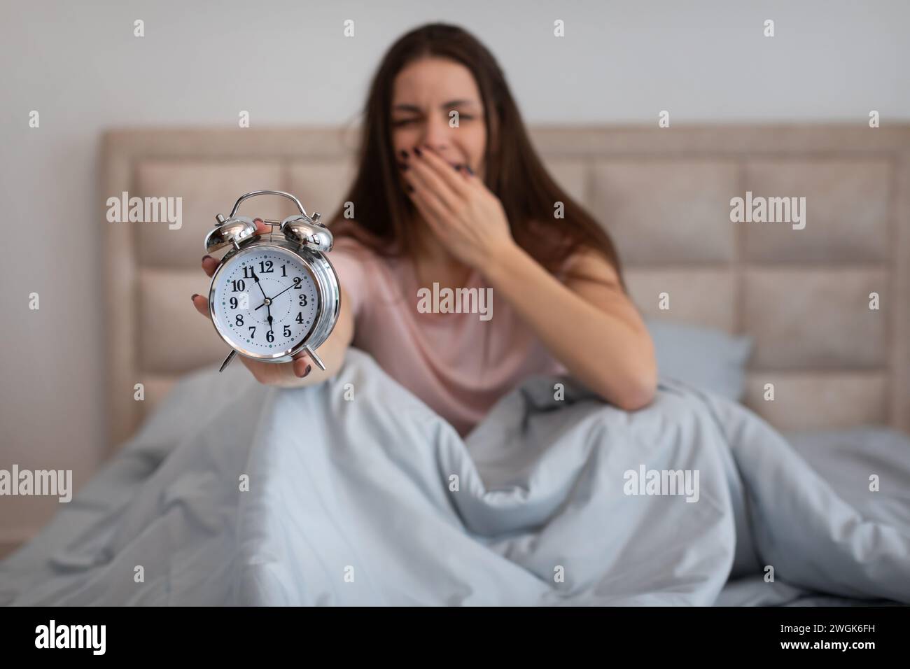 Sleepy woman in bed yawning, holding alarm clock in the morning Stock Photo