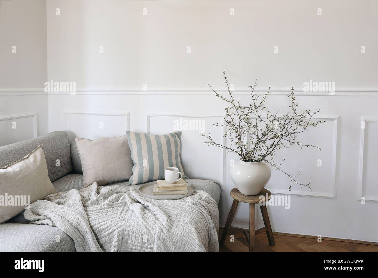 Modern spring scandinavian living room interior. Sofa with linen pale blue striped cushions and cup of coffee. Cherry plum blossoms in vase. Elegant Stock Photo