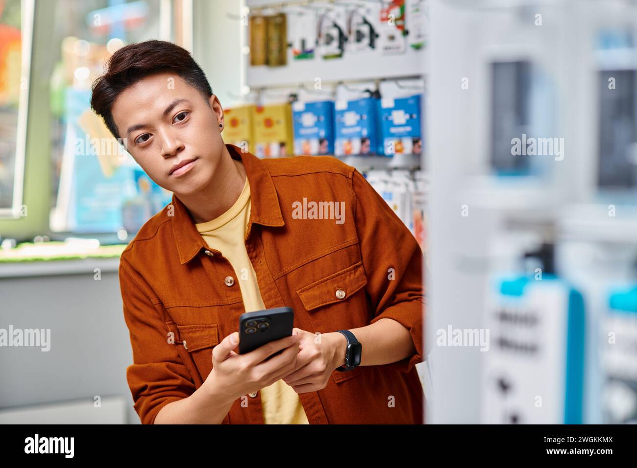 asian entrepreneur holding mobile phone and looking away in own electronics store, small business Stock Photo