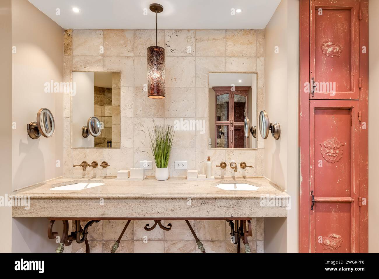 A Spanish style bathroom with brown stone tile walls, a light hanging over a granite countertop with gold mounted faucets, and a red rustic cabinet. Stock Photo