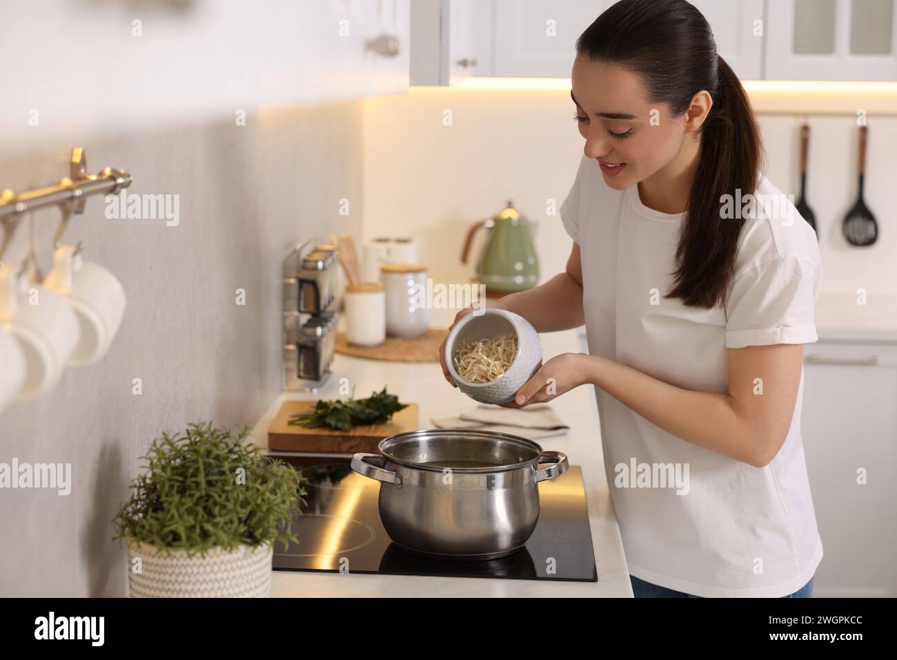 Smiling woman adding noodles into pot with soup in kitchen Stock Photo
