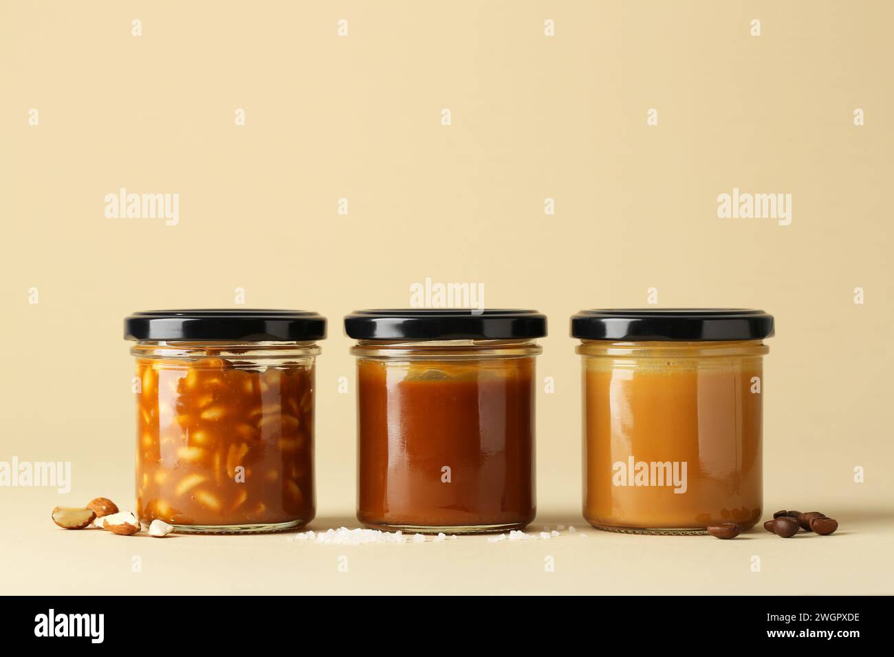 Tasty salted caramel in jars with peanuts, coffee beans and salt on pale yellow background Stock
