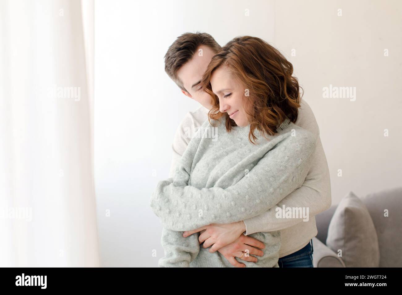 Loving young man embracing woman and kissing from behind in light living room at home. Romantic happy couple in casual clothes spending quality time t Stock Photo