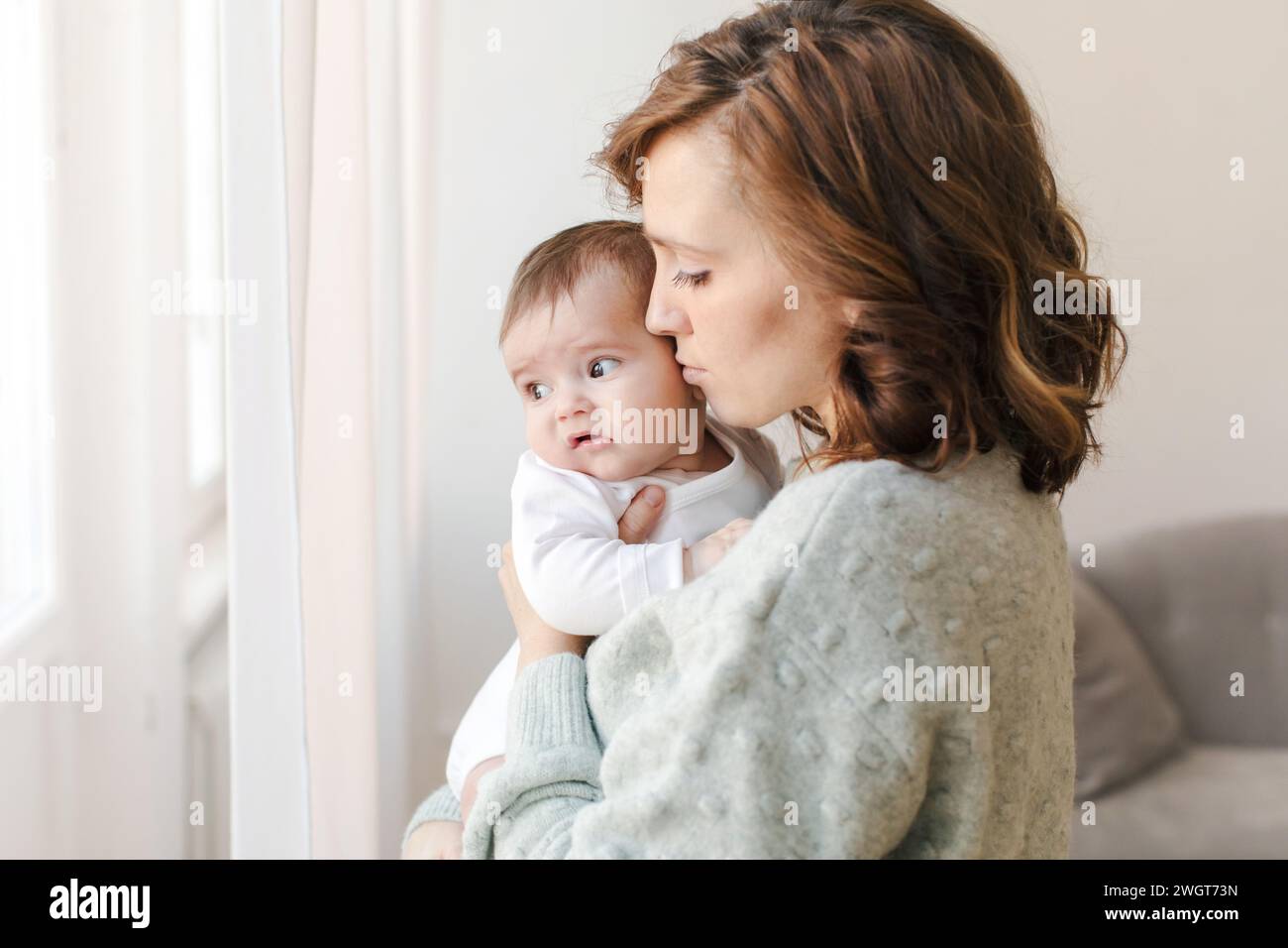 Side view of caring young mother in sweater holding and kissing adorable little baby while standing near window at home. Loving woman with infant spen Stock Photo