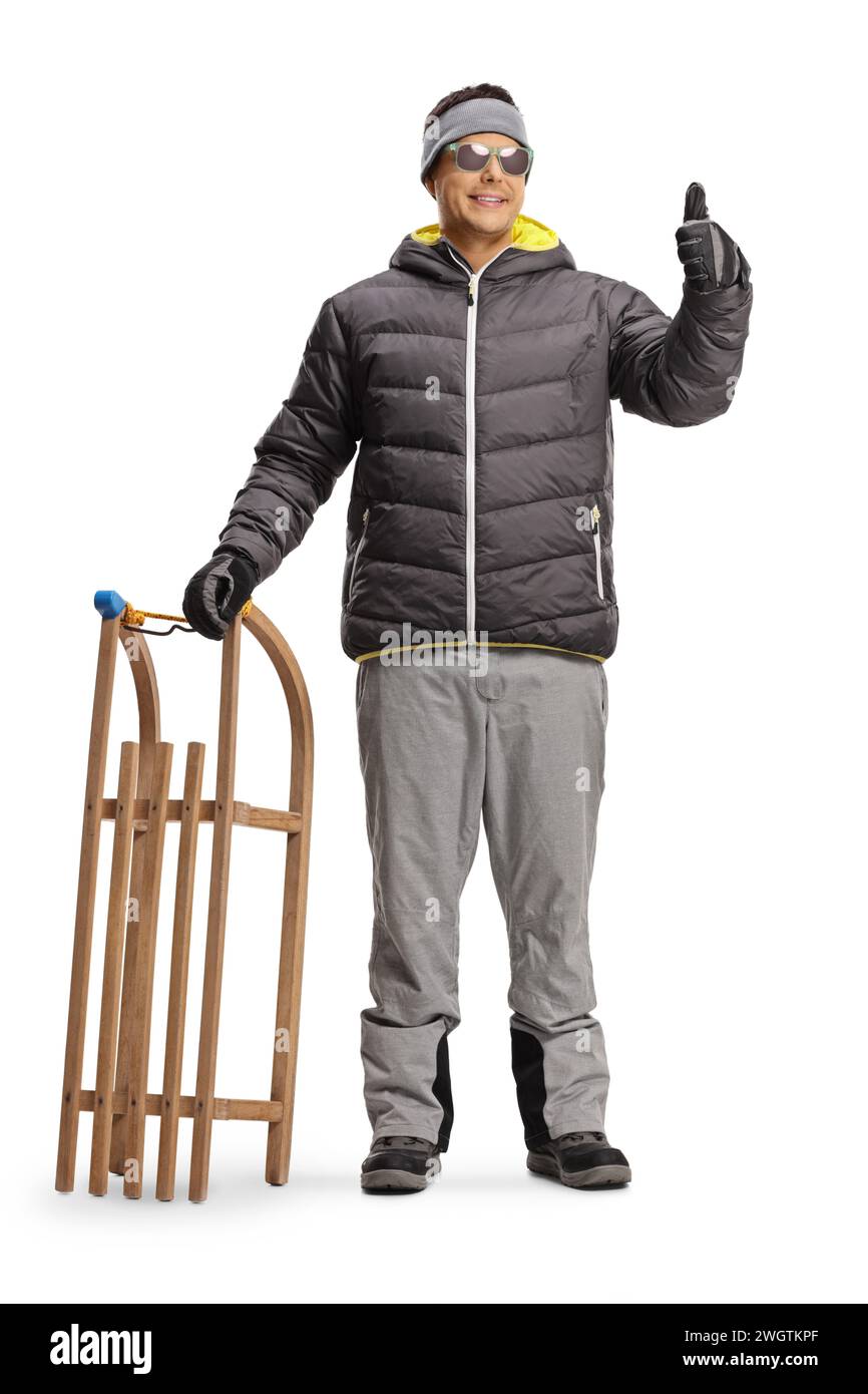 Young man in a winter jacket holding a wooden sled and gesturing thumbs up isolated on white background Stock Photo