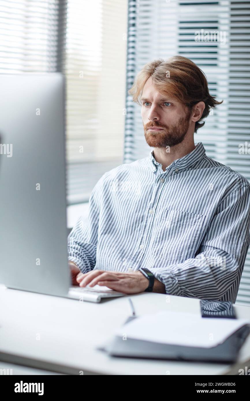 Graphic vertical portrait of bearded adult man using computer at workplace in IT office against window Stock Photo