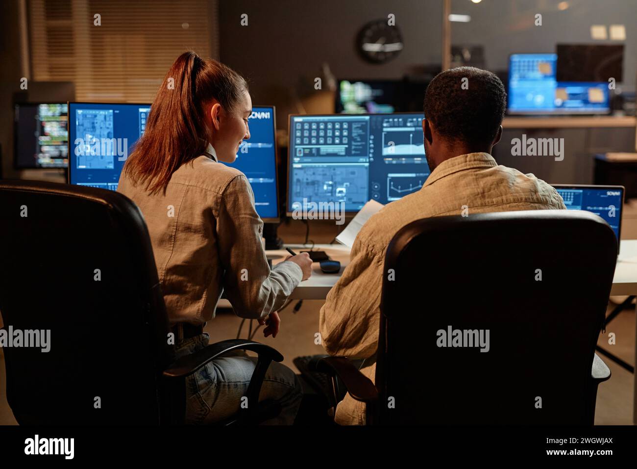 Back view at two IT developers man and woman looking at computer screen with data in dark office Stock Photo