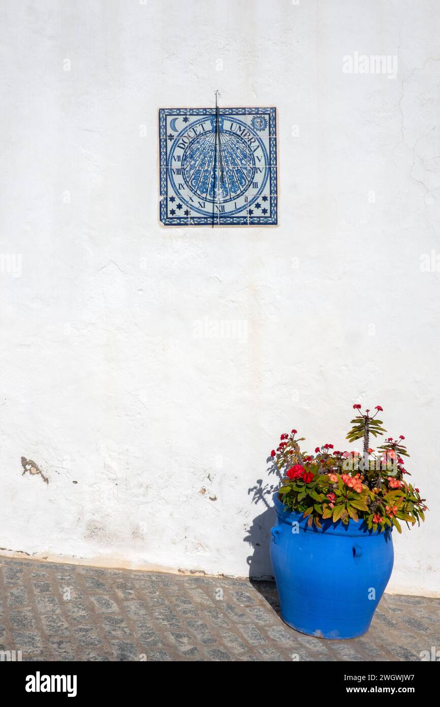 Sundial placed on a wall on a street in Vejer de la Frontera, Spain, next to a blue pot with flowers. Stock Photo