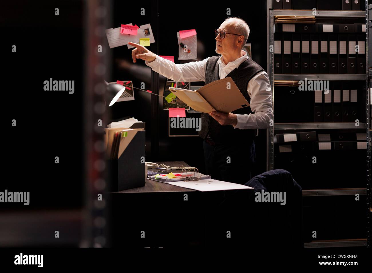 Senior police officer holding confidential files, looking at investigation board in arhive room. Elderly private detective working overhours at criminal case, checking mysterious suspect report Stock Photo