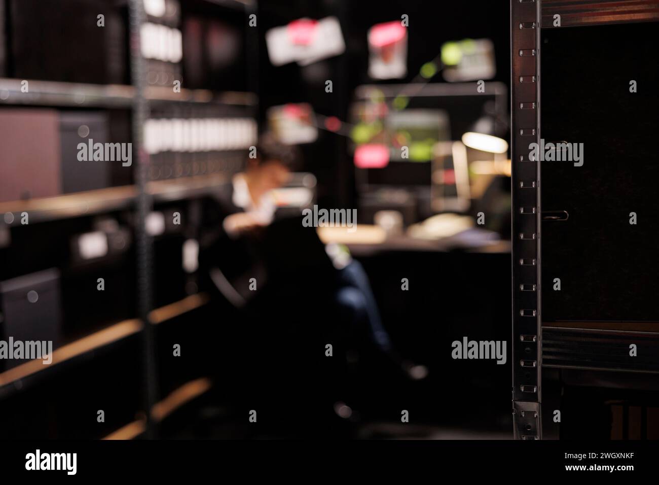 Selective focus of shelves full with evidence files, in background private investigator reading crime scene report. Police officer working late at night at missing person case in arhive room. Stock Photo