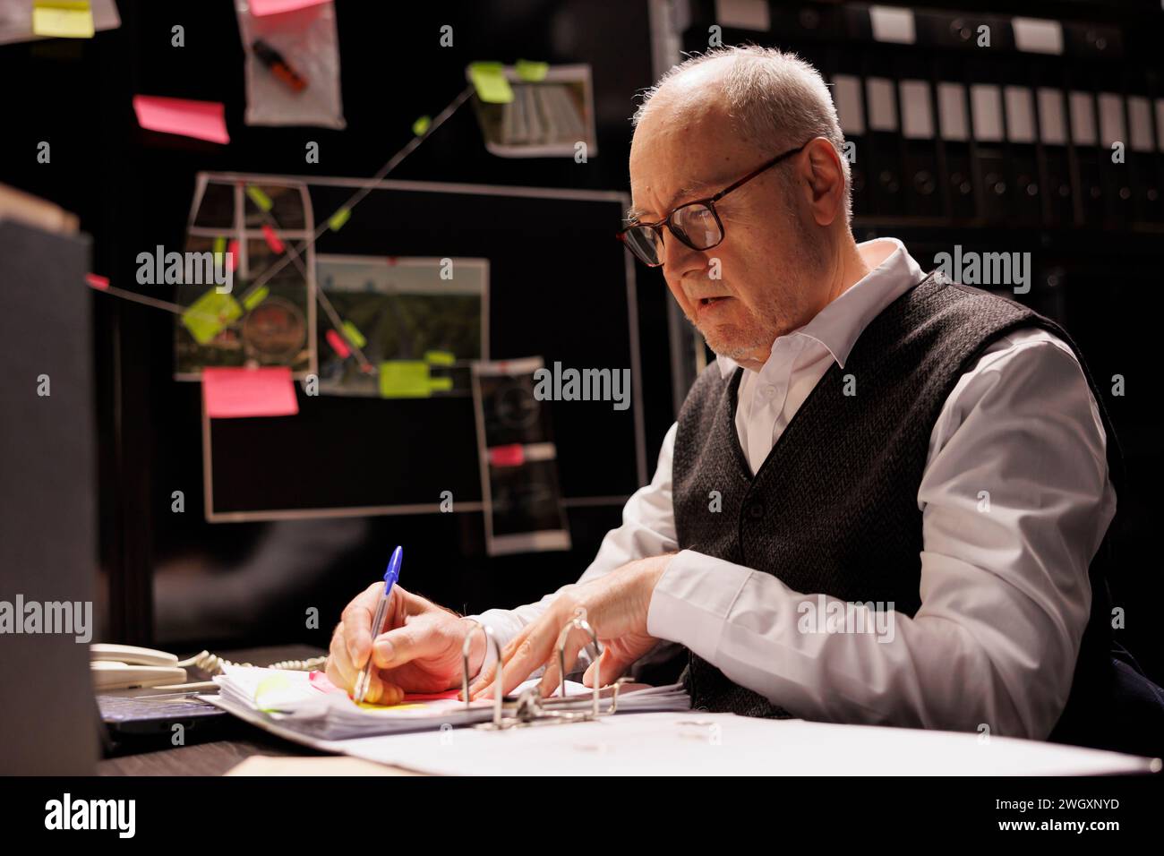 Tired elderly police officer sitting at desk in evidence room, analyzing late at night at criminology report. Overworked private detectives working at missing person case, checking victim files Stock Photo