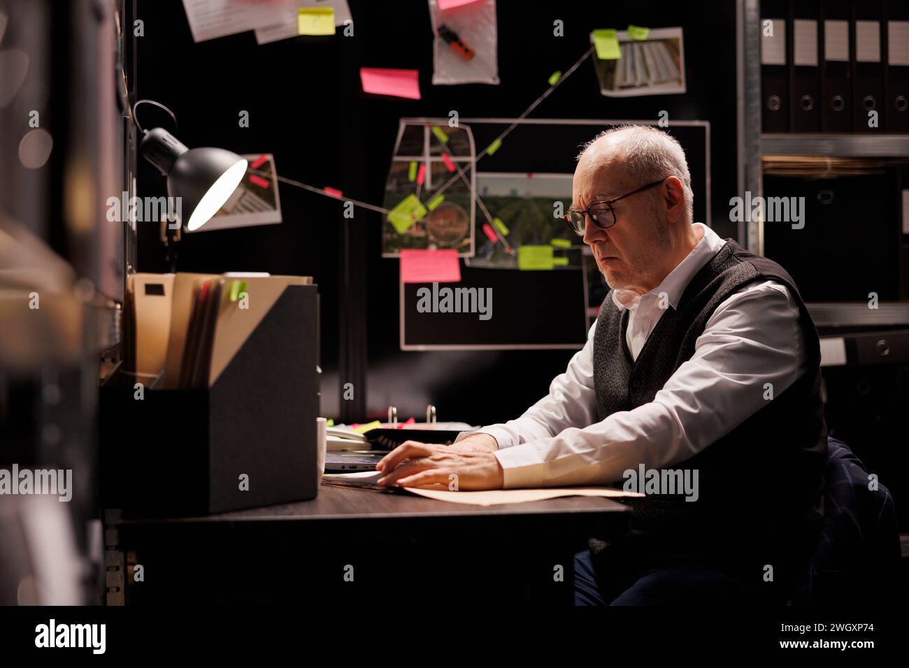 Tired elderly investigator analyzing criminology report, working overhours at criminal case in arhive room. Overworked private detective checking victim files, looking at crime scene evidence Stock Photo