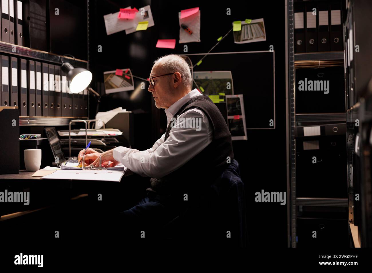 Elderly private detective writing criminal case report, working late at night at federal investigations in arhive room. Senior police officer analyzing crime scene evidence, checking suspect files Stock Photo