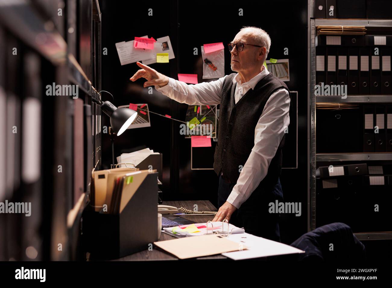 Elderly investigator working at criminal investigations case, analyzing evidence files in arhive room. Senior private detective checking criminology report, planning strategy to catch supect Stock Photo