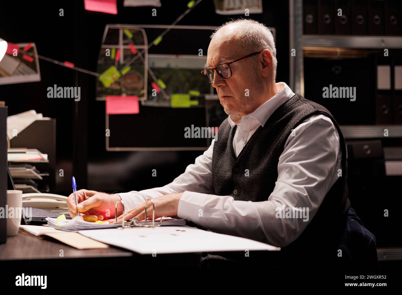 Senior investigator writing criminal case report, working overhours at federal investigations in arhive room. Tired elderly private detective analyzing crime scene evidence, checking suspect files Stock Photo
