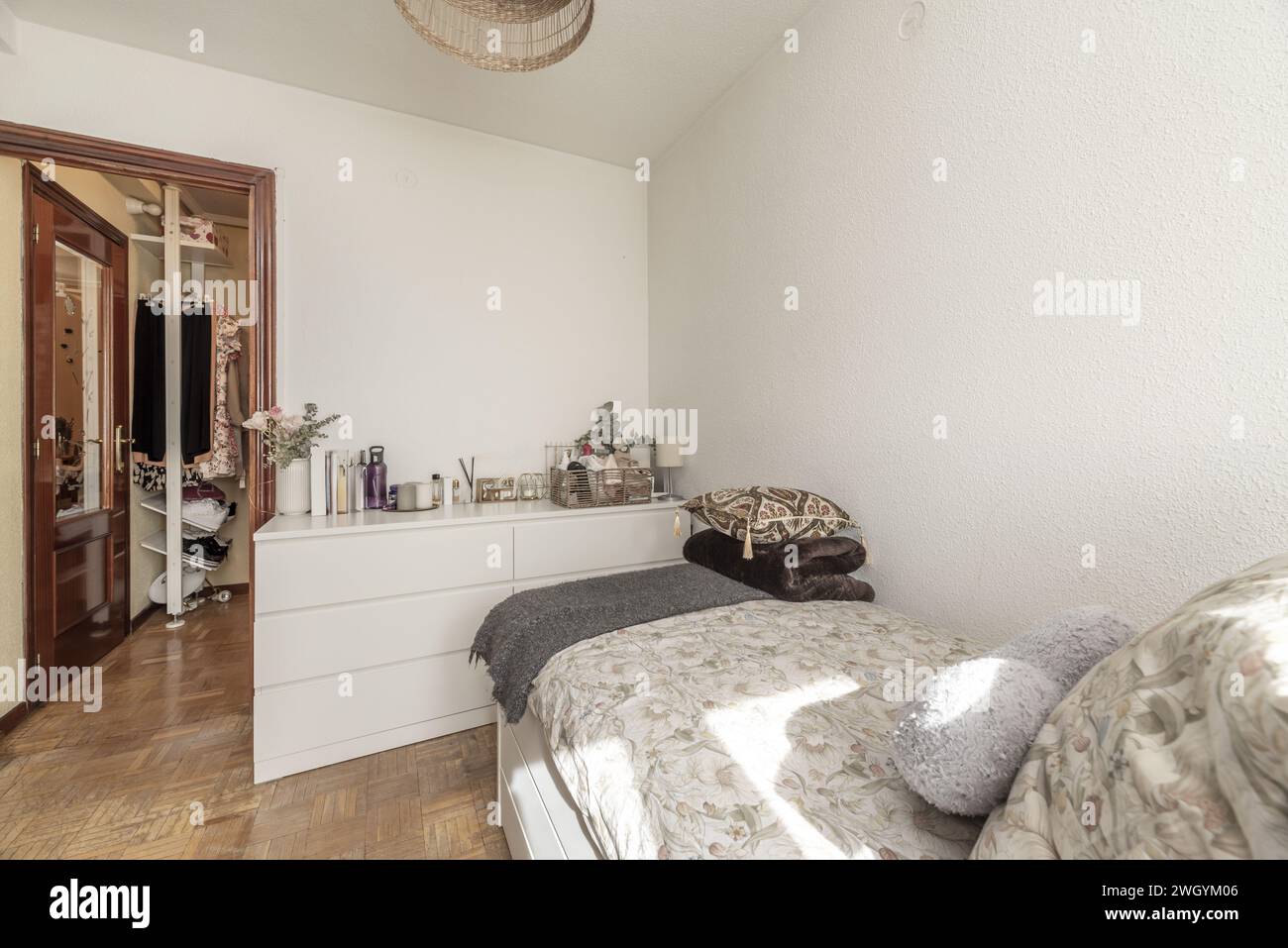 A youth bedroom with a single bed, aluminum radiator, clothes hanging on a shelf outside the room and a white dresser with many drawers Stock Photo